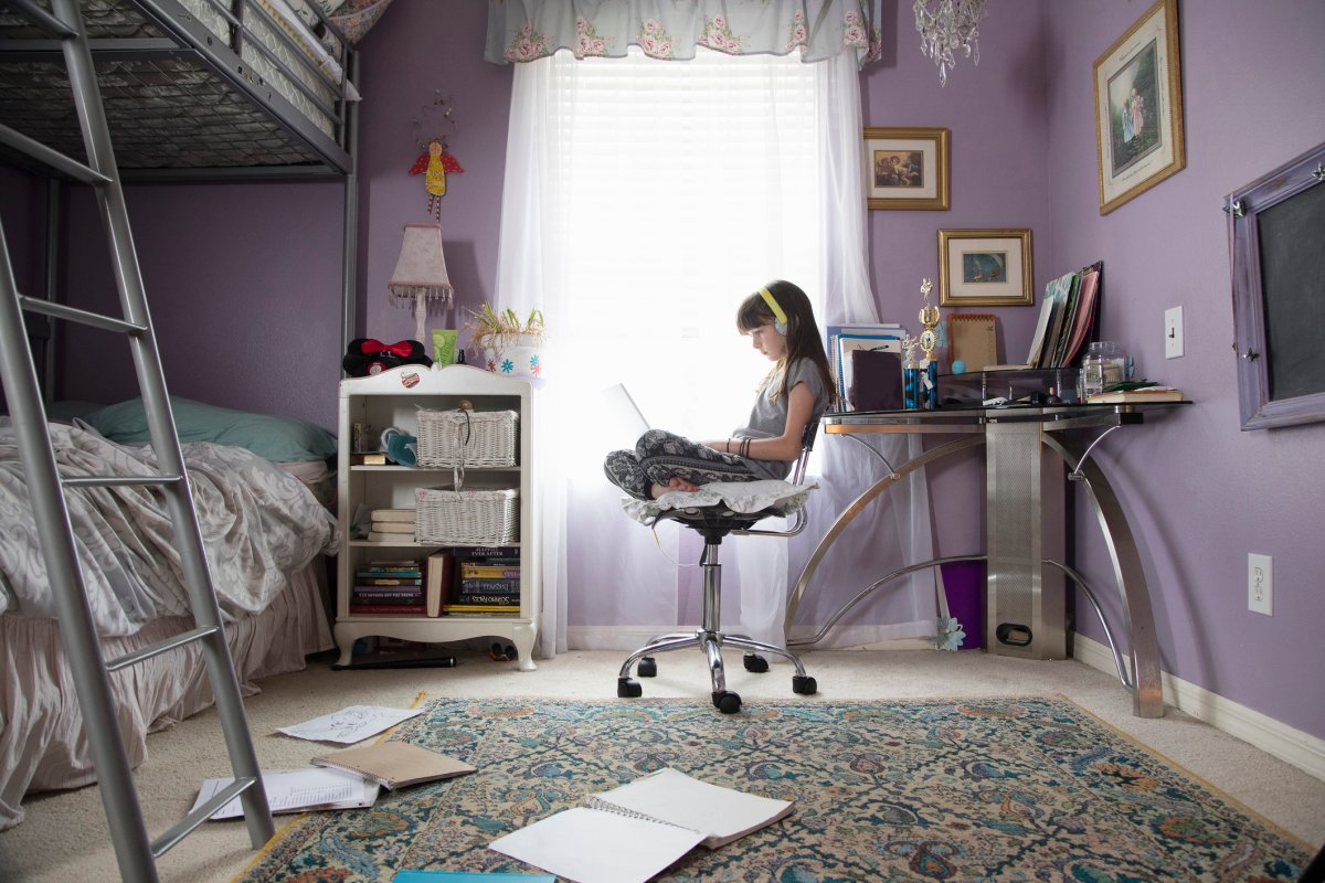 Girl sits in her bedroom on a computer chair with headphones on and looking down at her laptop in her lap. There are papers and some books on her floor and there is a bunkbed in her room